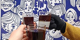 Foto: Drei Haende halten Tassen mit rotem Punsch vor einem blau-weissen Plakat hoch - auf dem Plakat zu erkennen das Wort Einsam