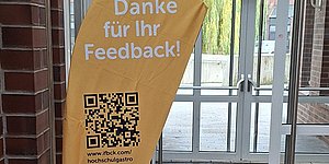 Foto: Gelbe Dropflag mit qr-Code, Aufdruck Danke fuer Ihr Feedback!, im Hintergrund Mauerwerk und Tuer mit Glasscheibe