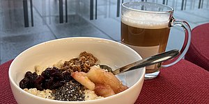 Foto: weisse Schale mit Porridge, Fruechten und Loeffel, sowie Glastasse mit Kaffee auf rotem Stoff vor Fenster