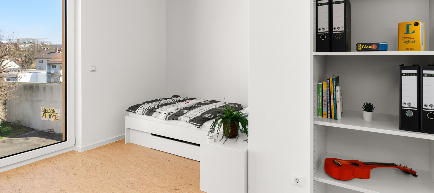 Einzelzimmer in WG mit bodentiefem Fenster. Zu sehen sind auch ein Bett sowie ein Bücherregal.
