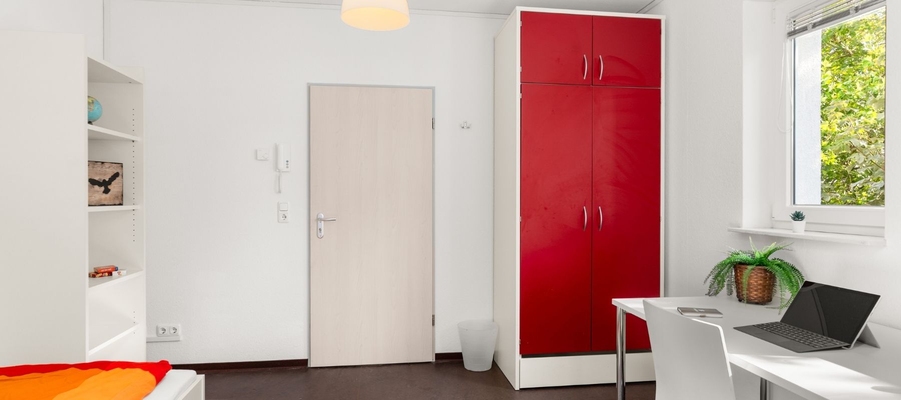 Foto eines Einzelapartments in einer WG. Zu sehen sind ein Schreibtisch, ein Bücherregal und ein Kleiderschrank.
