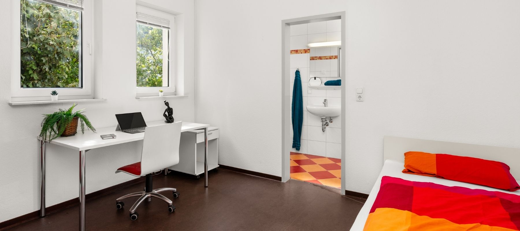 Foto eines Einzelapartments in einer WG. Zu sehen sind ein Schreibtisch, ein Bett sowie die Türe zum Bad des Apartments..