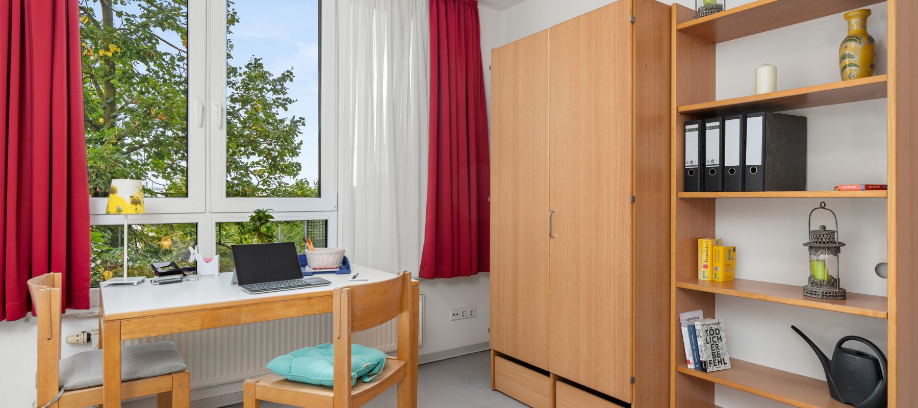 Foto eines Einzelapartments mit einem Schreibtisch am Fenster und einem Bücherregal.