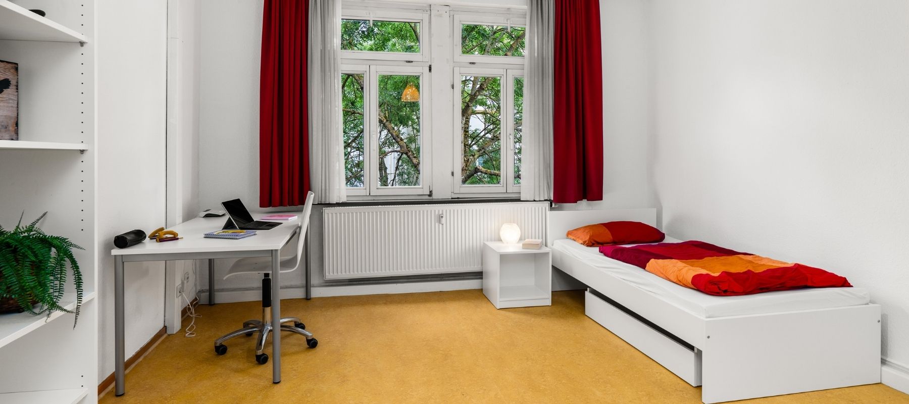 WG-Zimmer in der Hegelsbergstraße 20. Im Zimmer zu sehen sind ein Bett, große Fenster, ein Regal sowie ein Schreibtisch mit Stuhl.