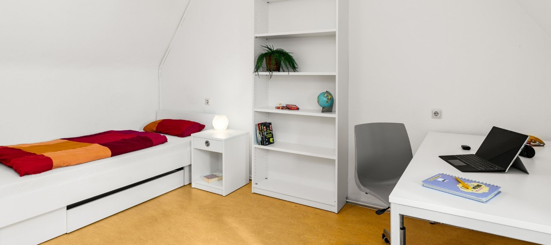WG-Zimmer in der Hegelsbergstraße 22. Zu sehen sind ein Bett mit Nachttisch, ein Regal sowie ein Schreibtisch mit Stuhl.