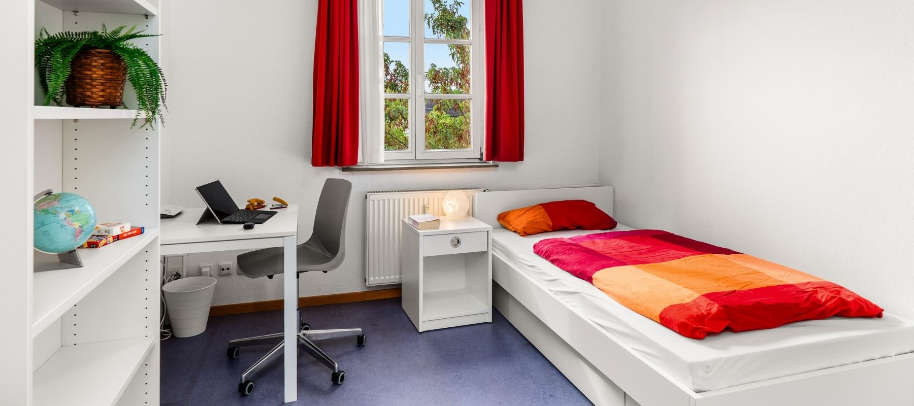 WG-Zimmer in der Hegelsbergstraße 24. Zu sehen sind ein Bett mit Nachttisch, ein Bücherregal sowie ein Schreibtisch mit Stuhl.