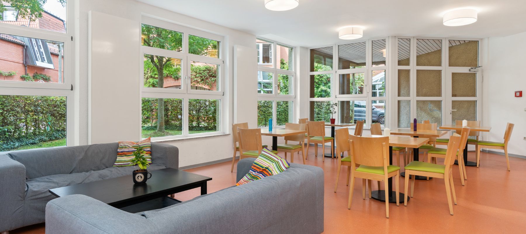 Gemeinschaftsraum des Wohnheims Nora-Platiel-Straße 7 mit Sofas sowie Tischen und Stühlen. Die großen Fenster geben den Blick auf Gebäude der Universität Kassel frei.