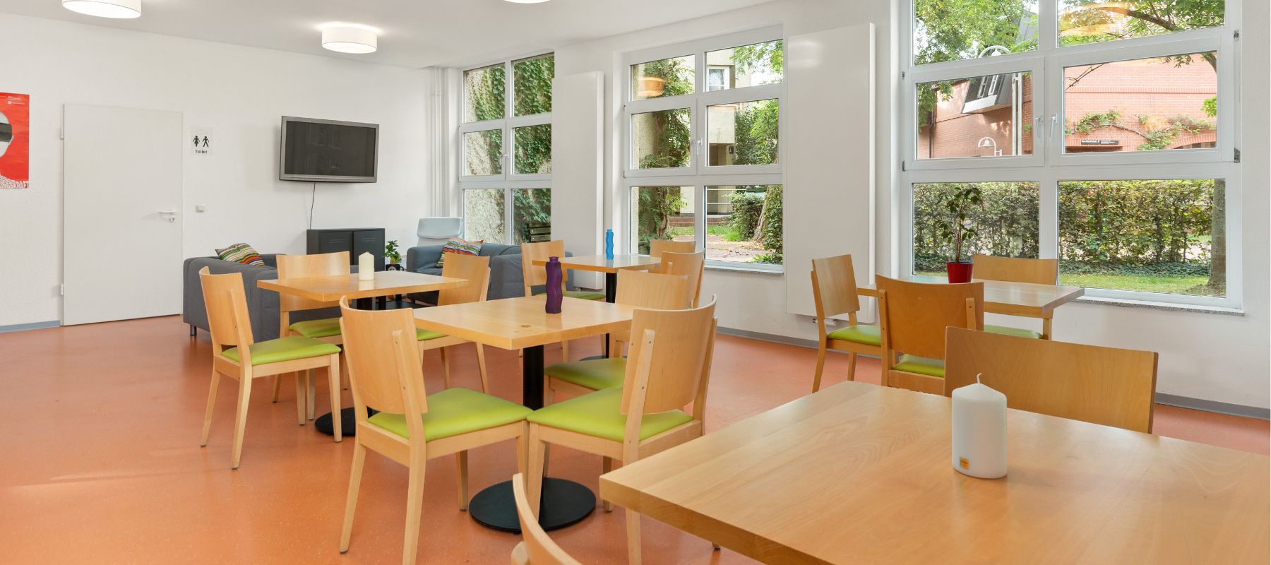 Gemeinschaftsraum im Wohnheim Nora-Platiel-Straße 7 mit Fernseher, Sofas sowie Tischen und Stühlen. An der hinteren Wand ist ein Toilettenschild zu erkennen.