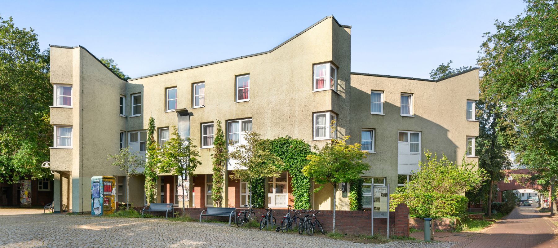 Eingang des Wohnheims Nora-Platiel-Straße 10. Davor ein Platz mit Kopfsteinpflaster sowie eine Mauer, Sitzbänke und abgestellte Fahrräder.