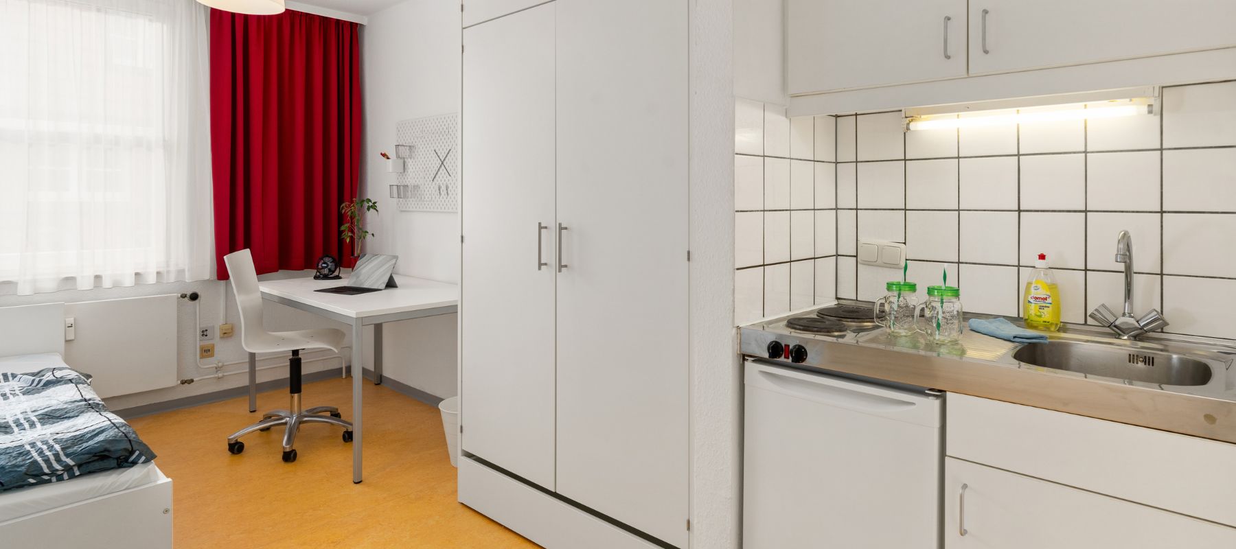 Einzelapartment mit Schreibtisch, Kleiderschrank, Küchenzeile und Bett.