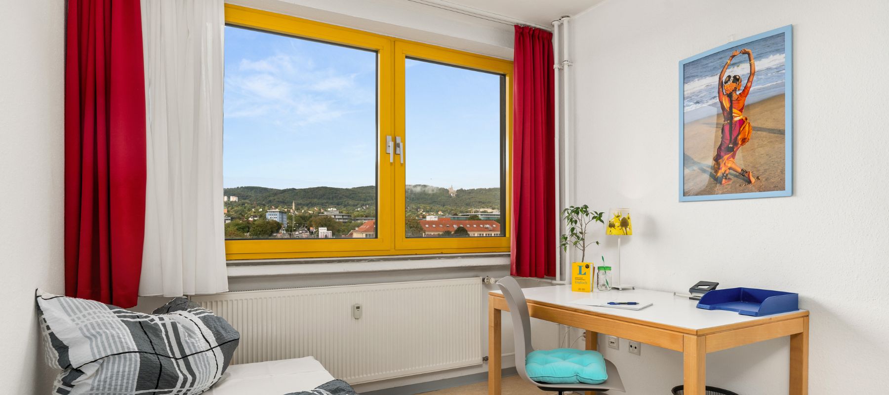 Einzelzimmer in einer WG. Zu sehen ist ein Schreibtisch am Fenster und der Ausschnitt eines Betts. Vom Fenster überblickt man die Stadt und vel Grün.