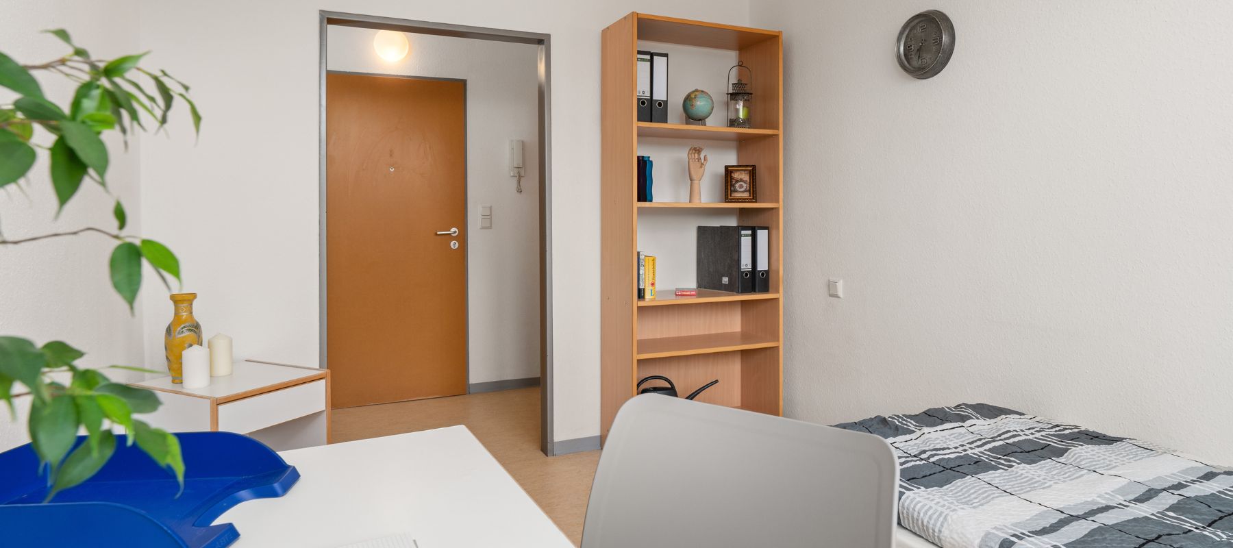 Einzelzimmer einer WG. Zu sehen sind ein Schreibtisch mit Stuhl, ein Bett, ein Bücherregal sowie die Zimmertüre.