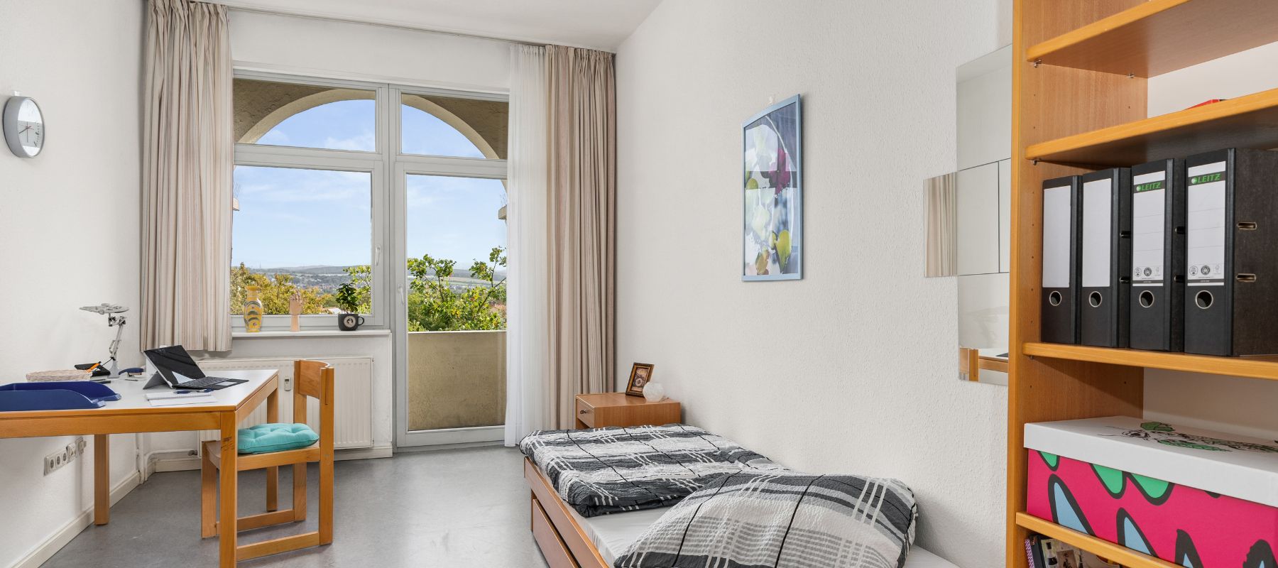 Blick in ein Einzelzimmer einer WG. Zu sehen sind große Fenster, ein Schreibtisch mit Stuhl, ein Bett mit Beistelltisch.