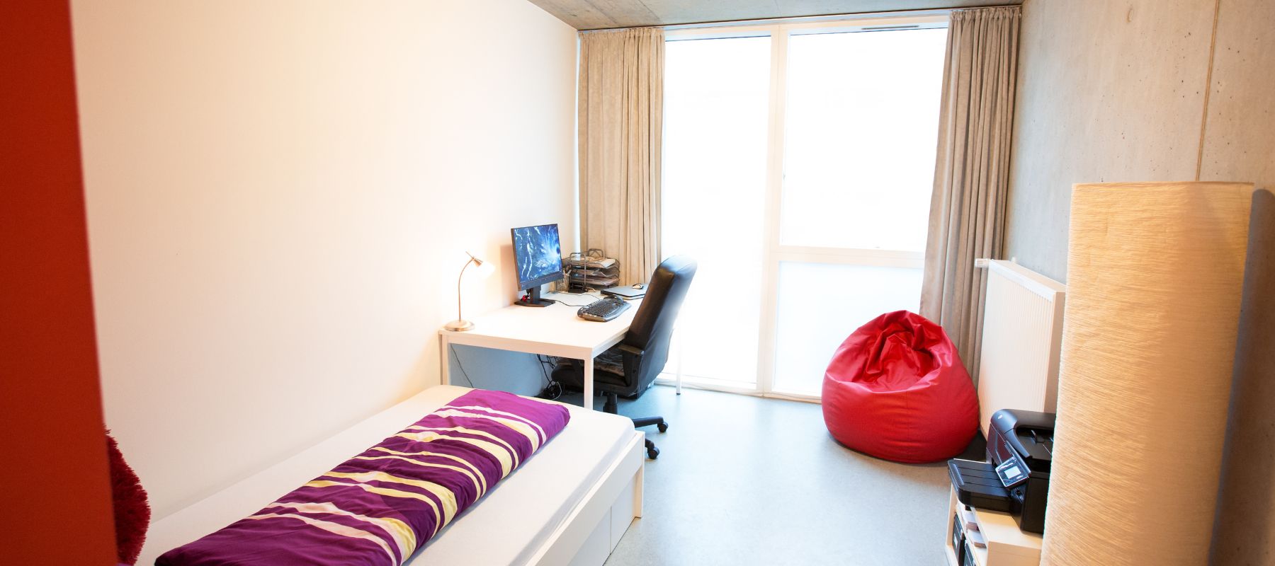 Blick in ein Einzelapartment. Zu sehen ist ein Bett, ein Schreibtisch mit Bürostuhl und ein Beistelltisch.