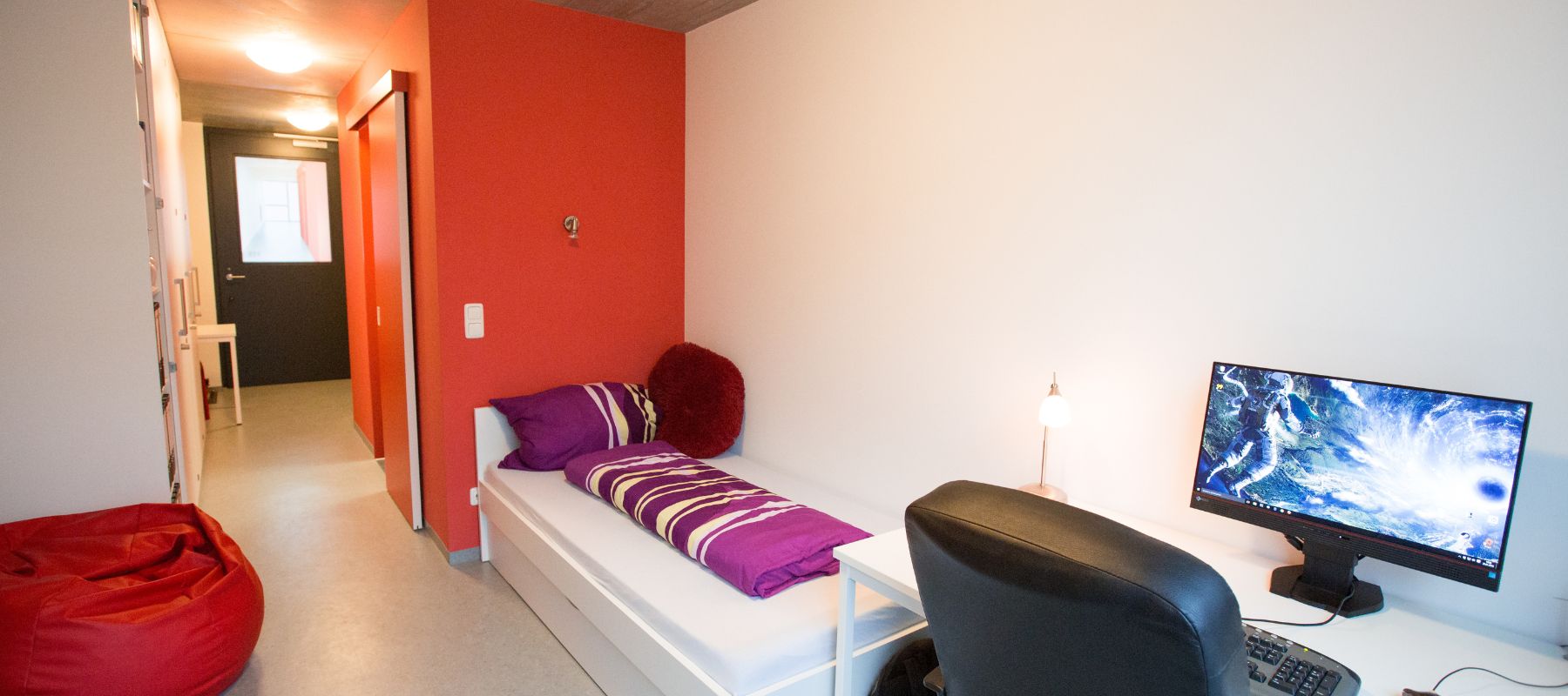 Blick in ein Einzelapartment. Zu sehen sind Schreibtisch, Bett und die Zimmertüre sowie Regale, Schränke und ein weiterer Raum, in dem sich das Badezimmer befindet.