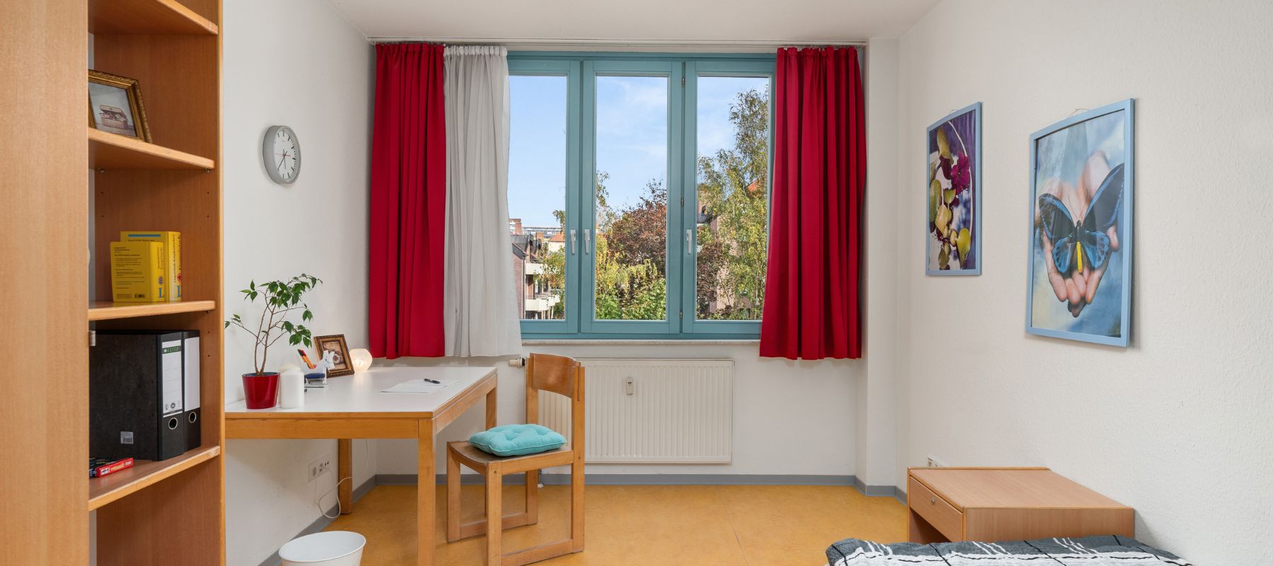 Blick in ein Einzelapartment. Zu sehen sind ein Bücherregal, ein Schreibtisch mit Stuhl sowie Bett und Beistelltisch.