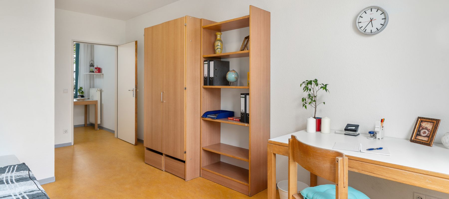 Blick in ein Einzelapartment. Zu sehen ist ein Regal, ein Schrank, ein Schreibtisch mit Stuhl sowie im Hintergrund die Küche des Apartments.