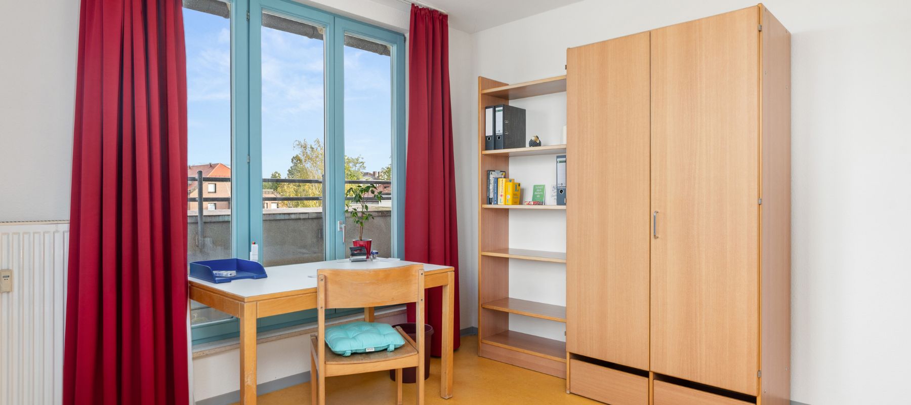 Einzelzimmer einer WG. Zu sehen sind Schreibtisch und Stuhl, die vor einem Fenster stehen, sowie ein Kleiderschrank und ein Regal an der Wand.
