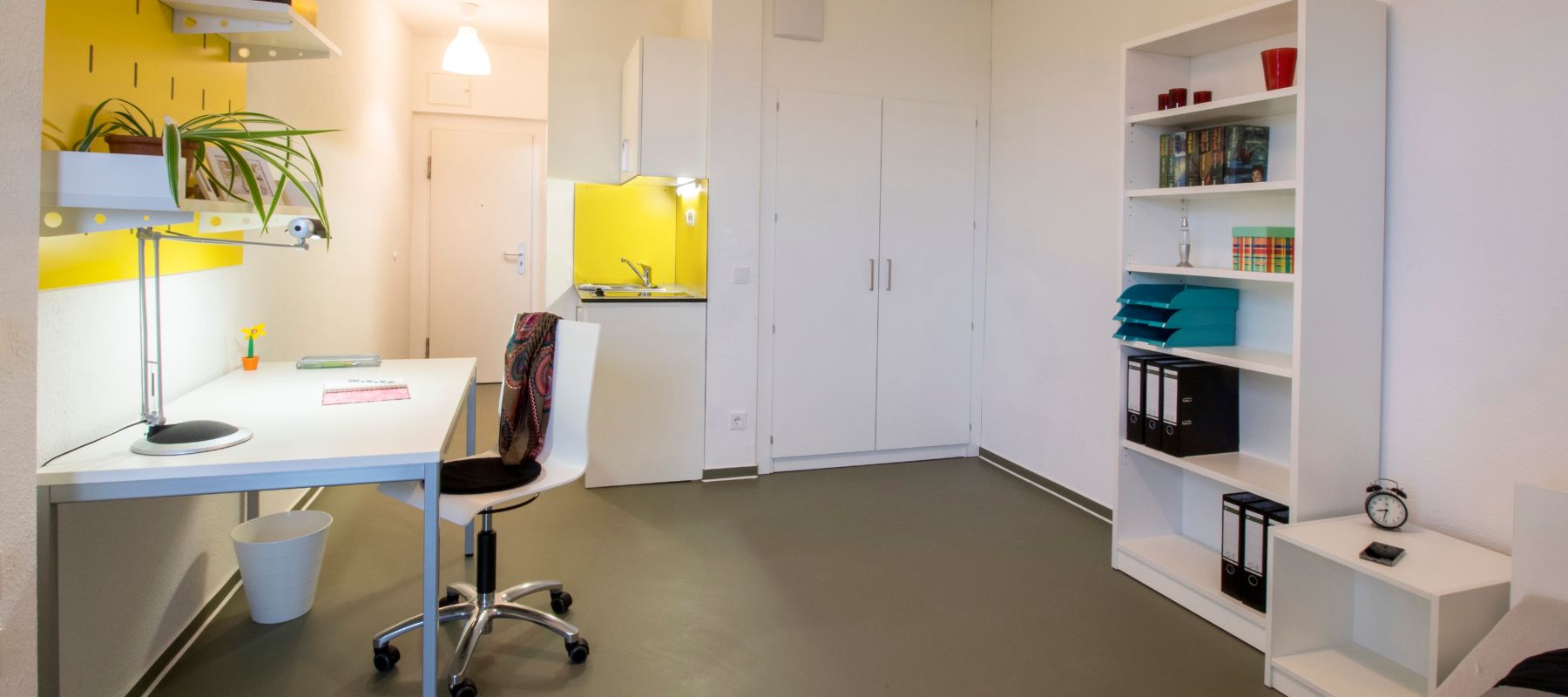 Einzelapartment im Wohnheim. Zu sehen ist ein Schreibtisch, der an der Wand steht, dazu ein Stuhl. Dahinter steht ein Bücherregal. Im Hintergrund die Küchenzeile, ein Kleiderschrank und die Türe zum Apartment. 