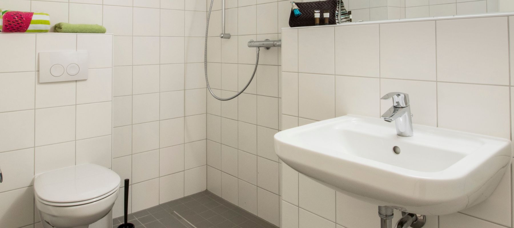 Bad eines Einzelapartments mit weißen Fließen. Zu sehen ist eine ebenerdige Dusche, eine Toilette sowie ein Waschbecken.