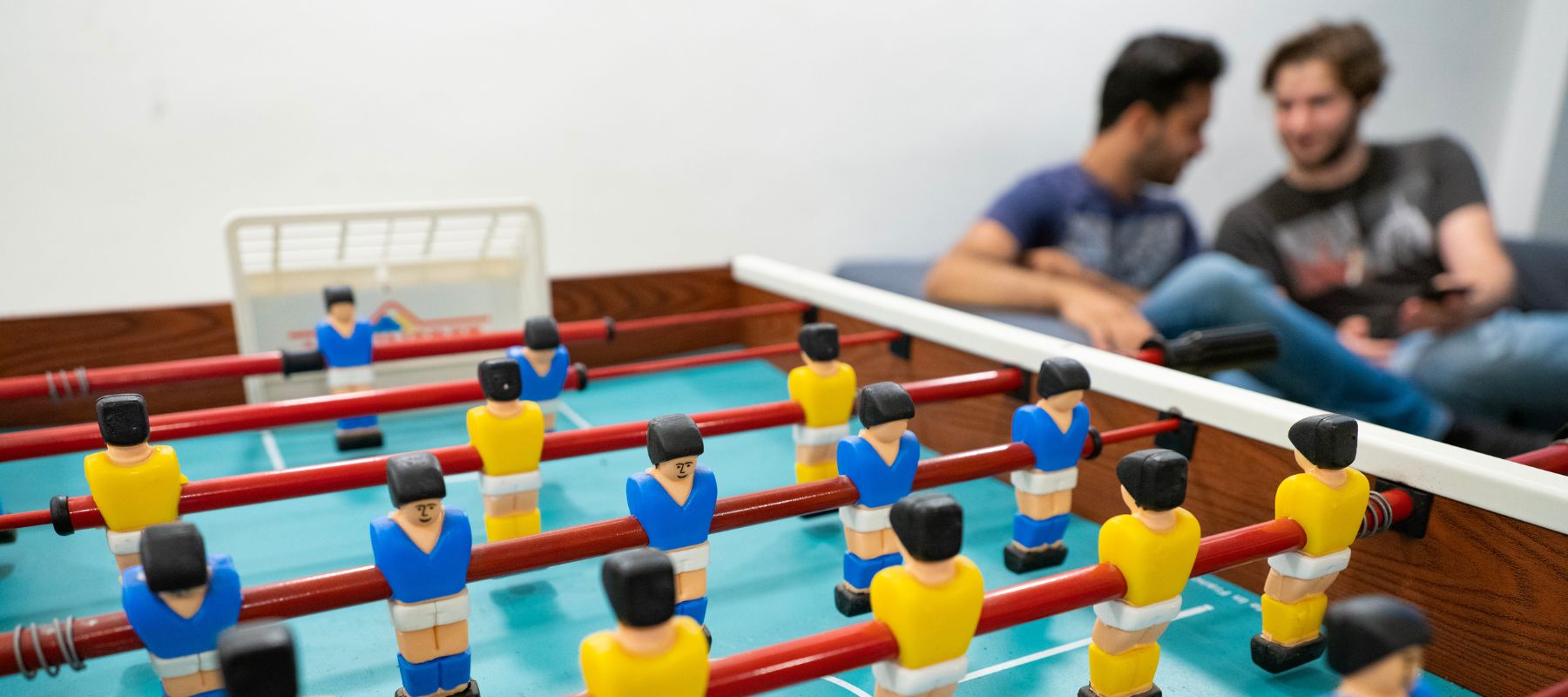 Im Vordergrund steht der Kickertisch im Gemeinschaftsraum des Wohnheims. Im Hintergrund sitzen zwei Studierende auf einem Sofa.