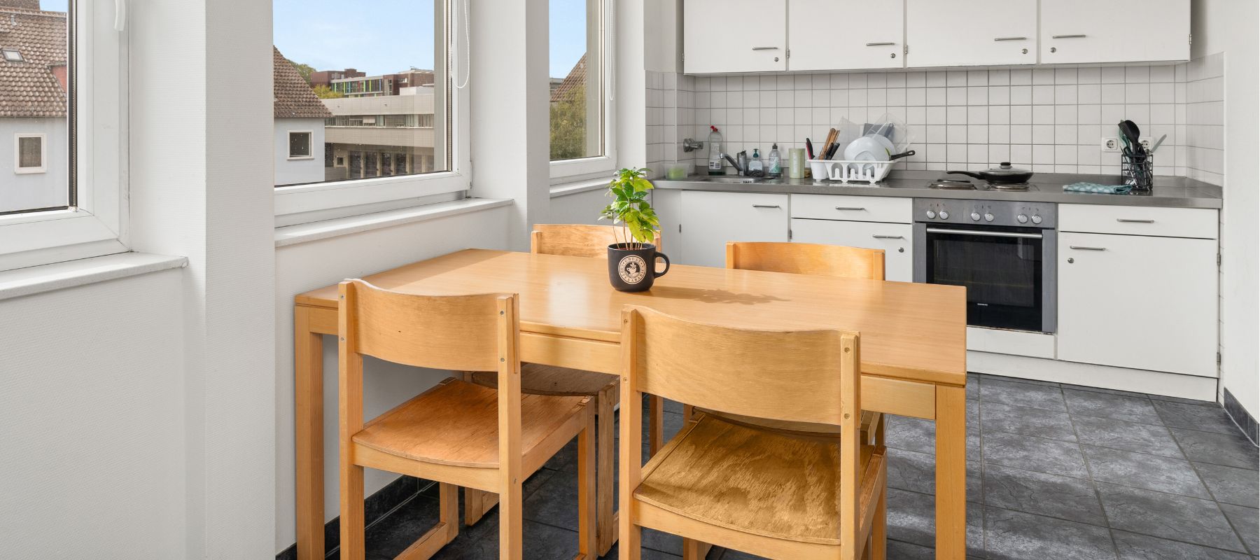 Eine WG-Küche. Im Vordergrund der Esstisch mit vier Stühlen, im Hintergrund die Küchenzeile mit Backofen, Herdplatten, Spüle und Schränken.