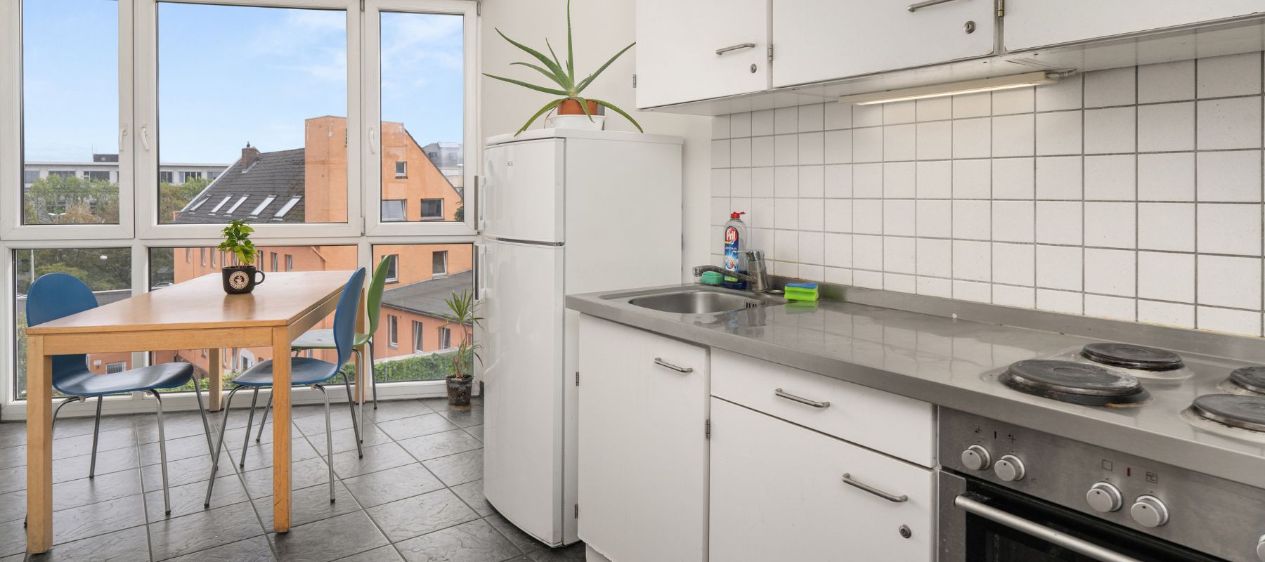 WG-Küche. Der Esstisch steht vor bodentiefen Fenstern, die Küchenzeile hat einen Kühlschrank, einen Backofen, vier Herplatten und eine große Spüle.