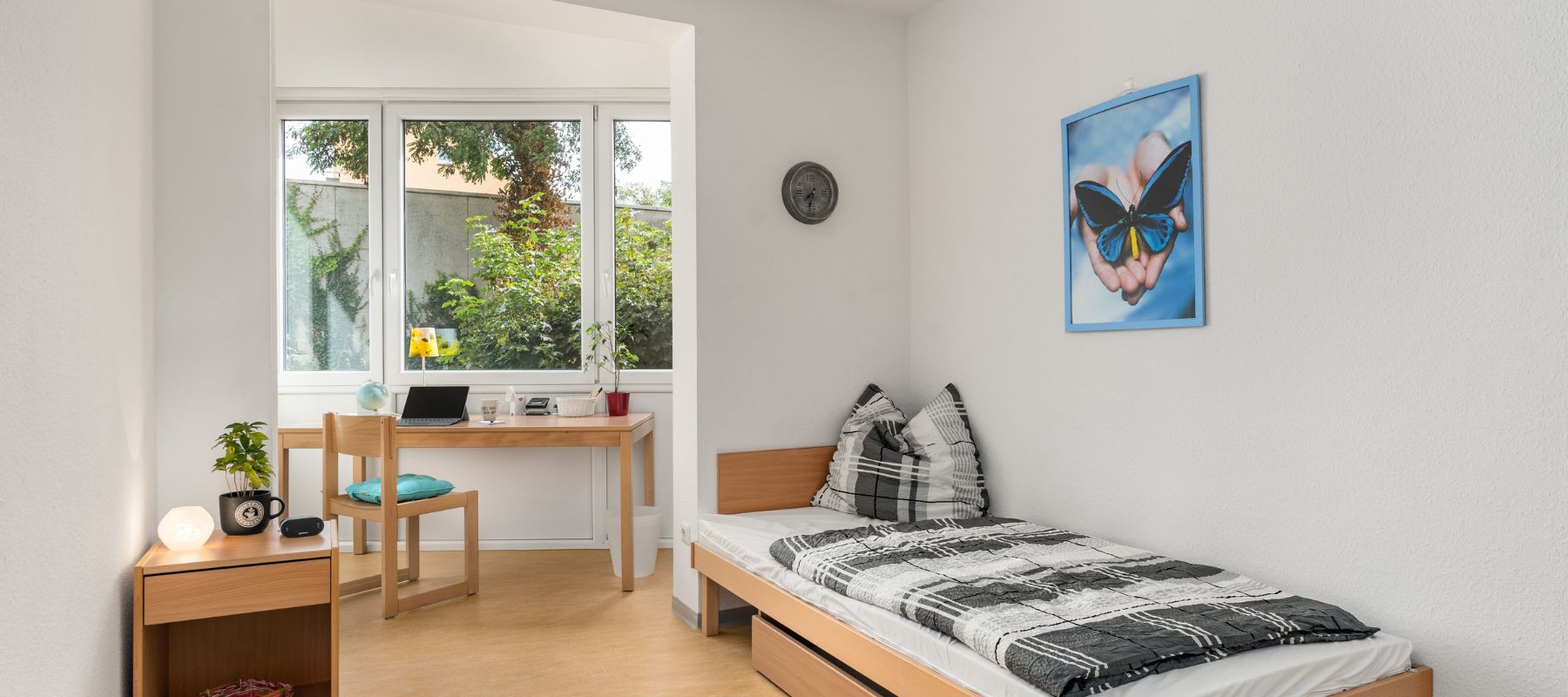 Einzelapartment im Wohnheim. Zu sehen ist ein Schreibtisch mit Stuhl, der vor einem Fenster steht, ein Bett sowie ein Nachtschränkchen.