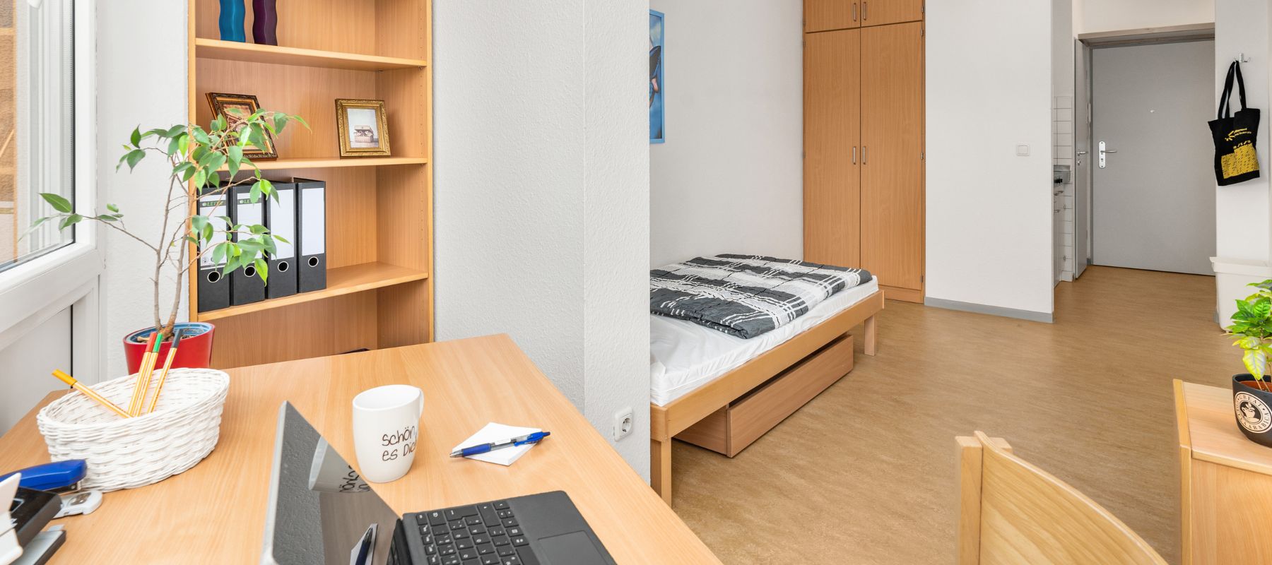 Einzelapartment im Wohnheim mit Schreibtisch und Stuhl. Im Hintergrund sieht man außerdem ein Bett, einen Kleiderschrank sowie die Türe zum Hausflur.