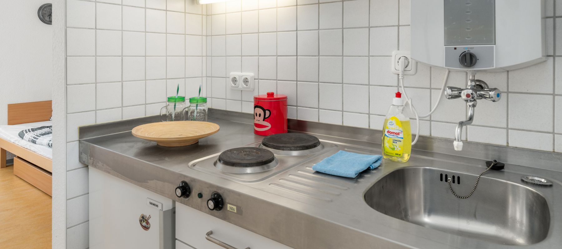 Kleine Küchenzeile im Einzelapartment mit Kühlschrank, Spüle und zwei Herdplatten.