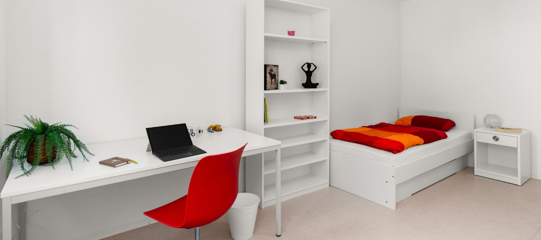 Foto eines Zimmeres in einem Doppelapartment. Zu sehen sind ein Schreibtisch mit Stuhl, ein Bücherregal und ein Bett.