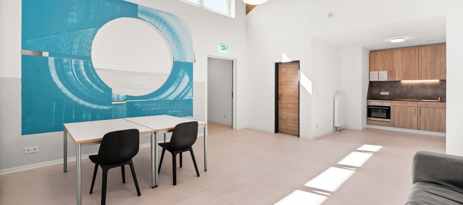 Foto von der Gemeinschaftsküche im 2. OG. Zu sehen sind Küchenzeile, Stühle, ein Küchentisch und ein großes Bild an der Wand.