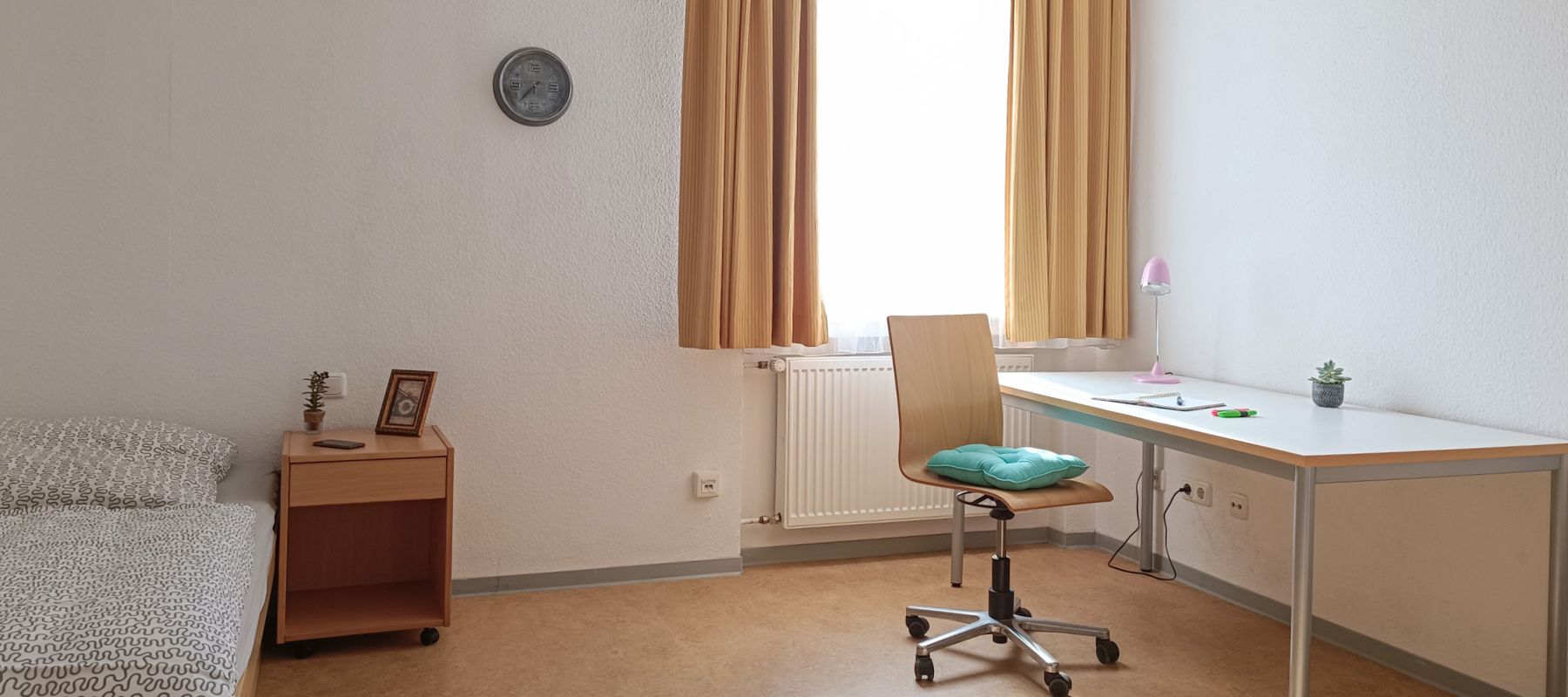 Innenansicht eines Einzelapartments. Zu sehen ist ein Schreibtisch mit Stuhl sowie ein Bett mit Beistelltisch.