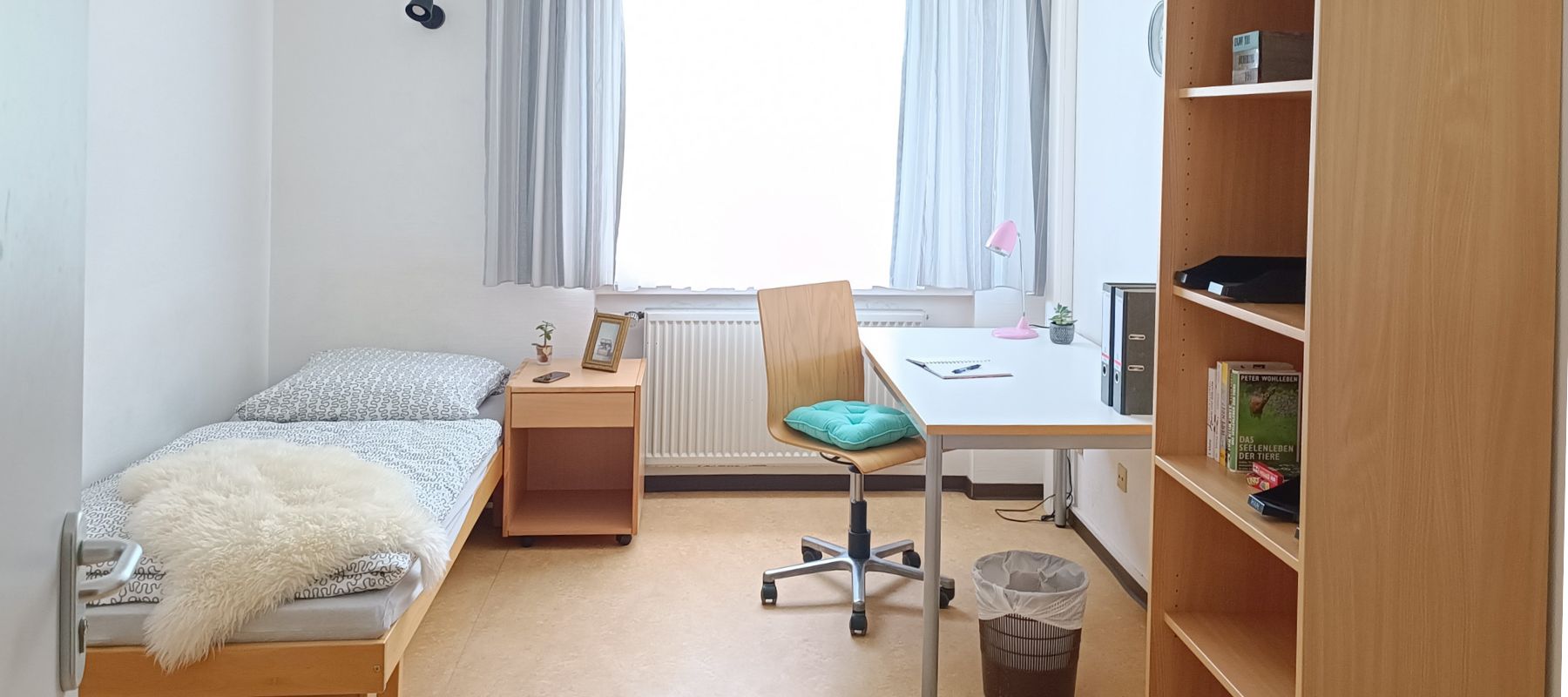 Einzelzimmer in einer WG. Zu sehen ist ein Bett mit Beistelltisch, ein Schreibtisch mit Stuhl sowie ein Bücherregal.