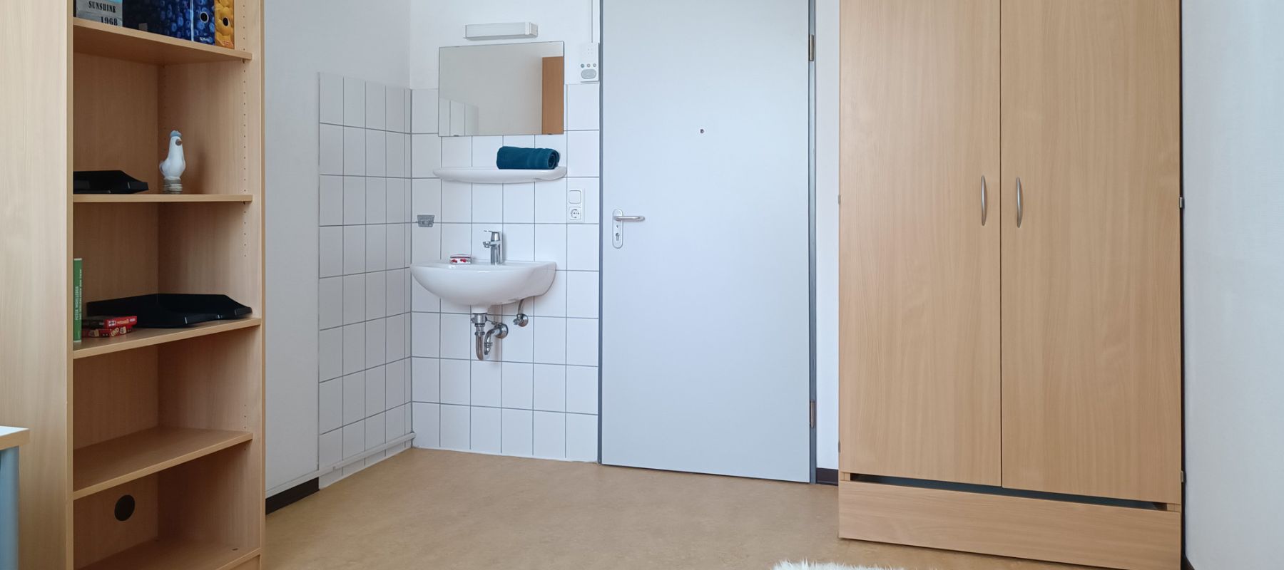 Einzelzimmer in einer WG mit Kleiderschrank, Waschbecken und Bücherregal.