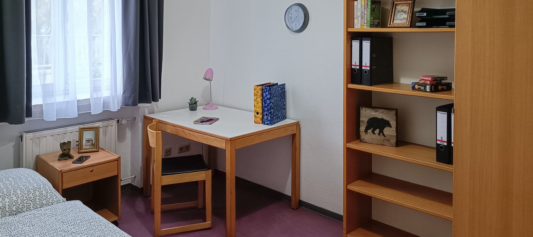 Zimmer im Doppelapartment mit Schreibtisch und Stuhl, Bett und Beistelltisch sowie einem Bücherregal,